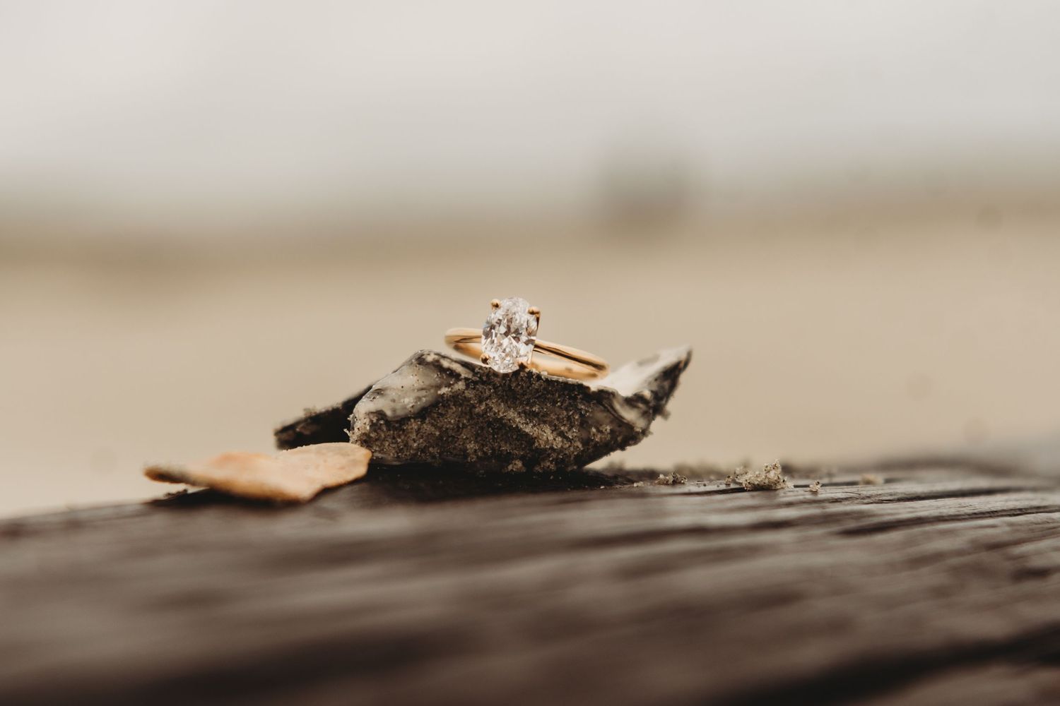 Gold diamond engagement ring resting on a rock on a wooden surface at a beach.