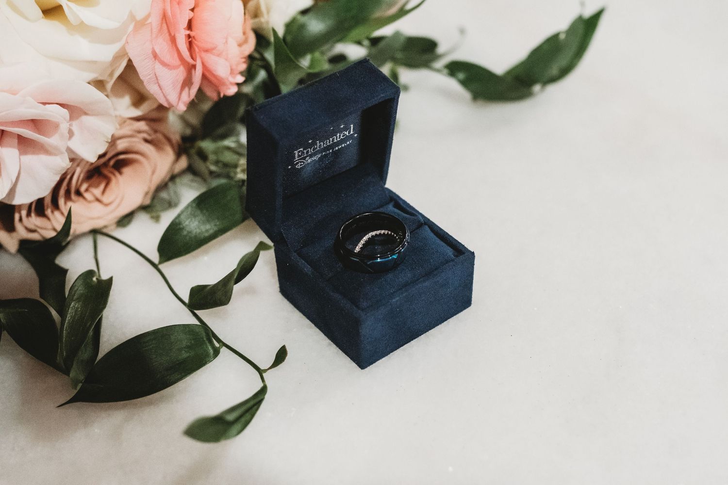 Wedding rings in navy blue box with pink roses and greenery on white background.