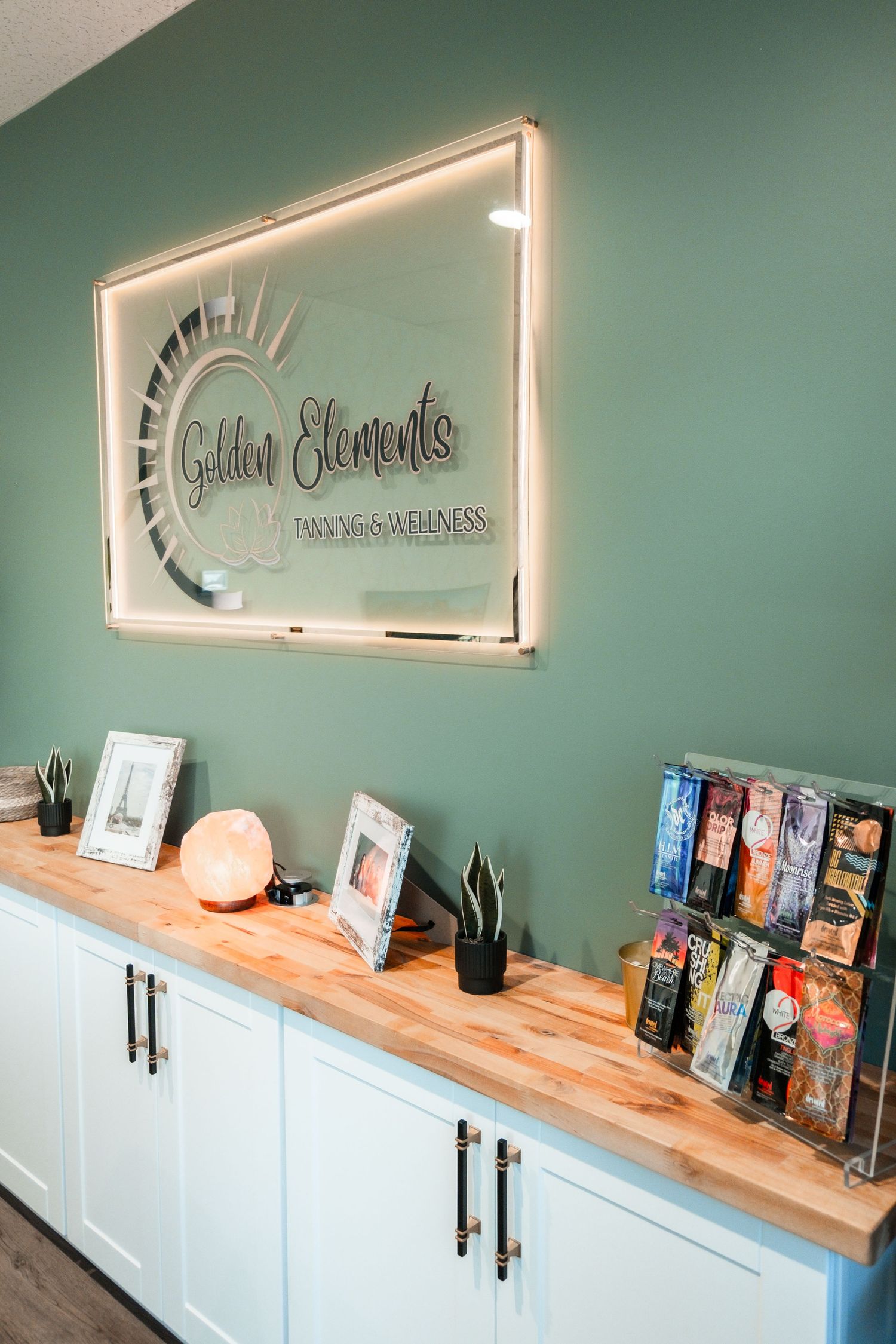 Golden Elements Tanning & Wellness reception desk with illuminated logo sign on sage green wall.