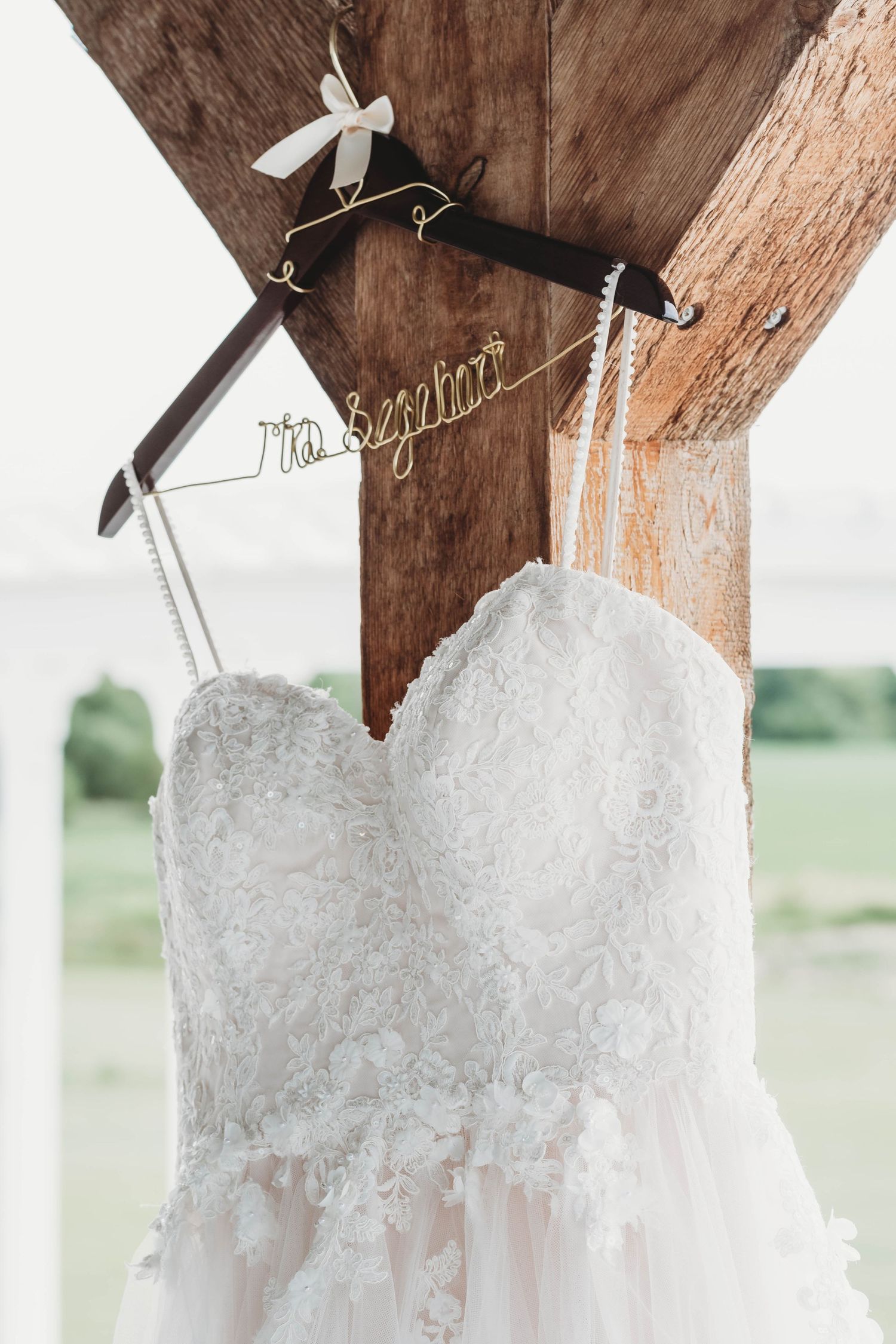 White lace wedding dress hangs elegantly on wooden beam with personalized hanger.