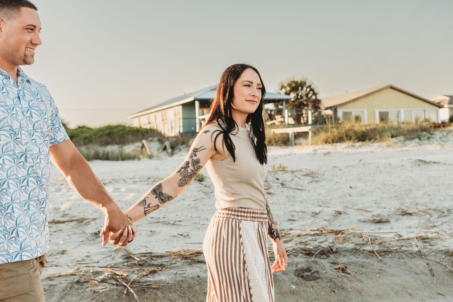Couple holding hands on a sandy beach at sunset, woman with tattoos wearing striped pants and a white top.