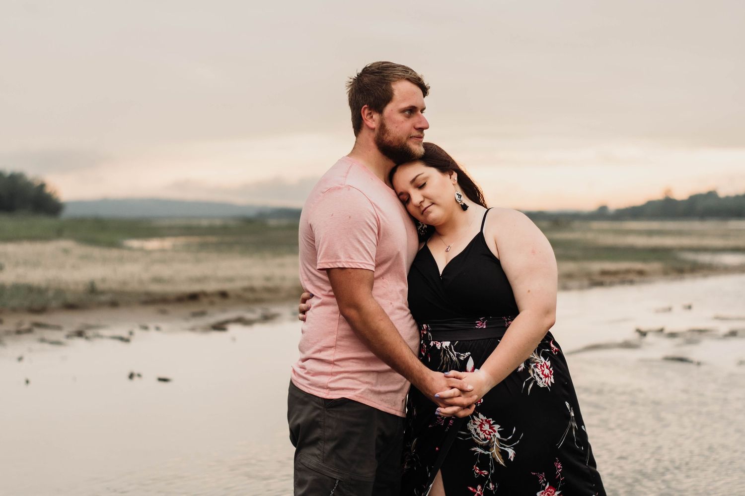 Couple embracing at sunset near a serene waterway, woman in floral dress leaning on mans shoulder.