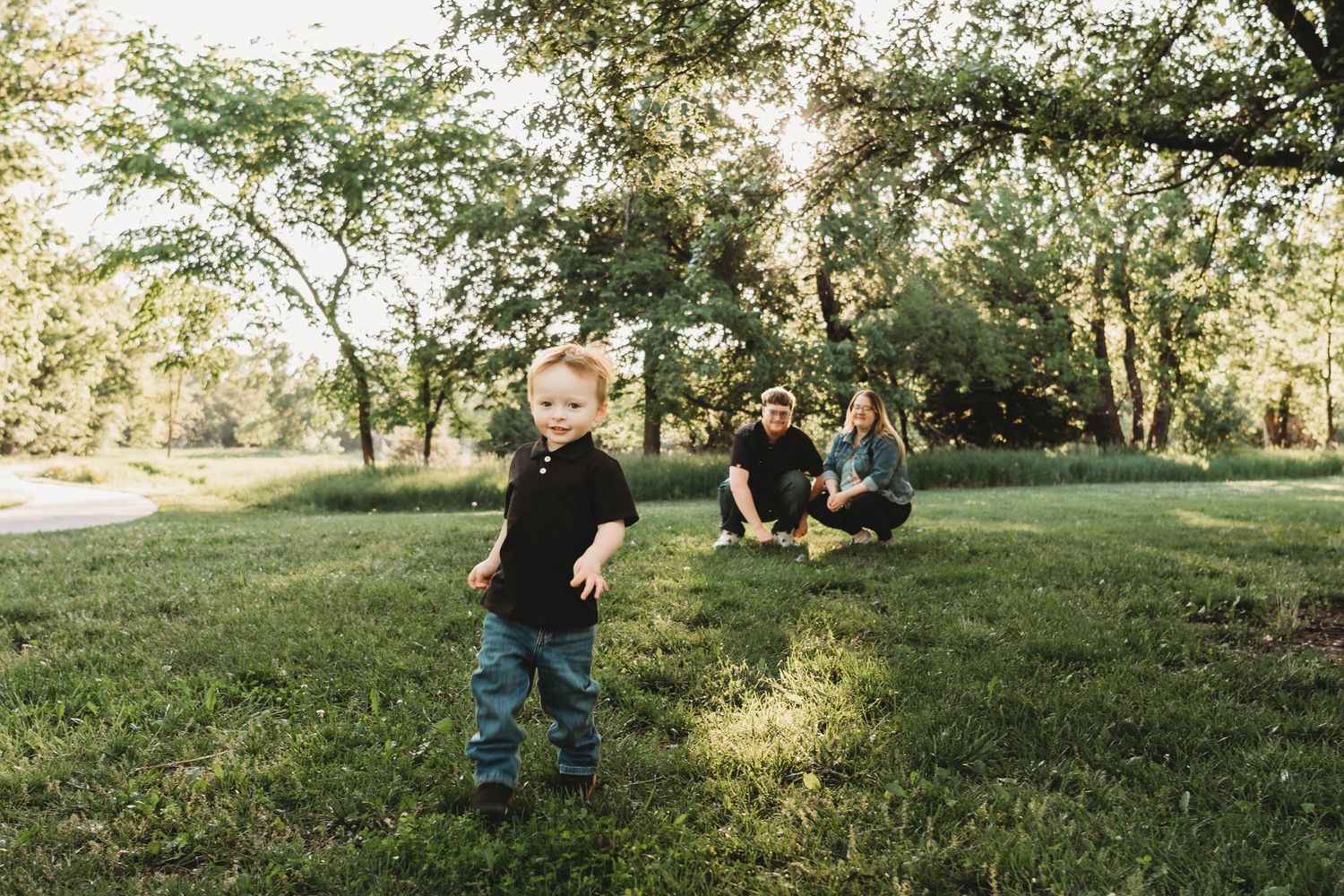 A toddler runs on green grass in a sunny park while two adults sit and watch in the background.