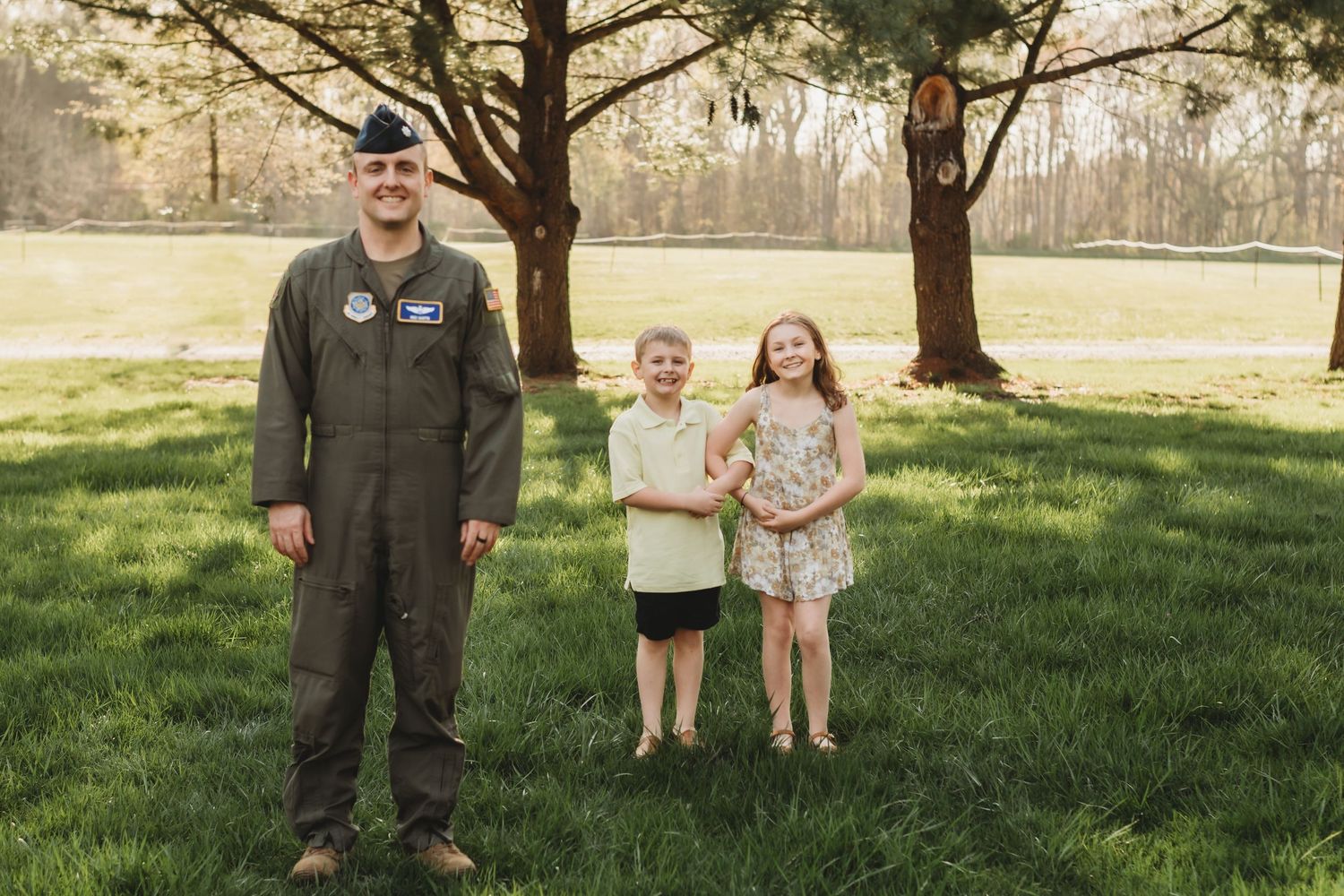 A person in a military flight suit stands in a grassy field while others watch in the background.