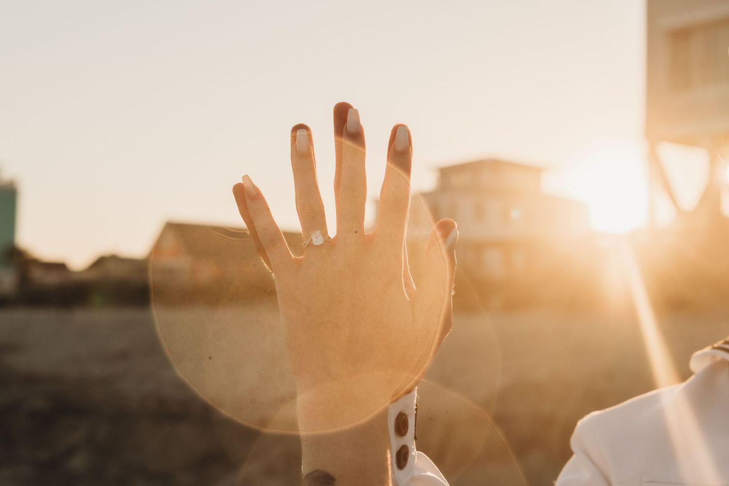 A hand raised against a golden sunset sky with lens flare, wearing a ring, with buildings in the background.