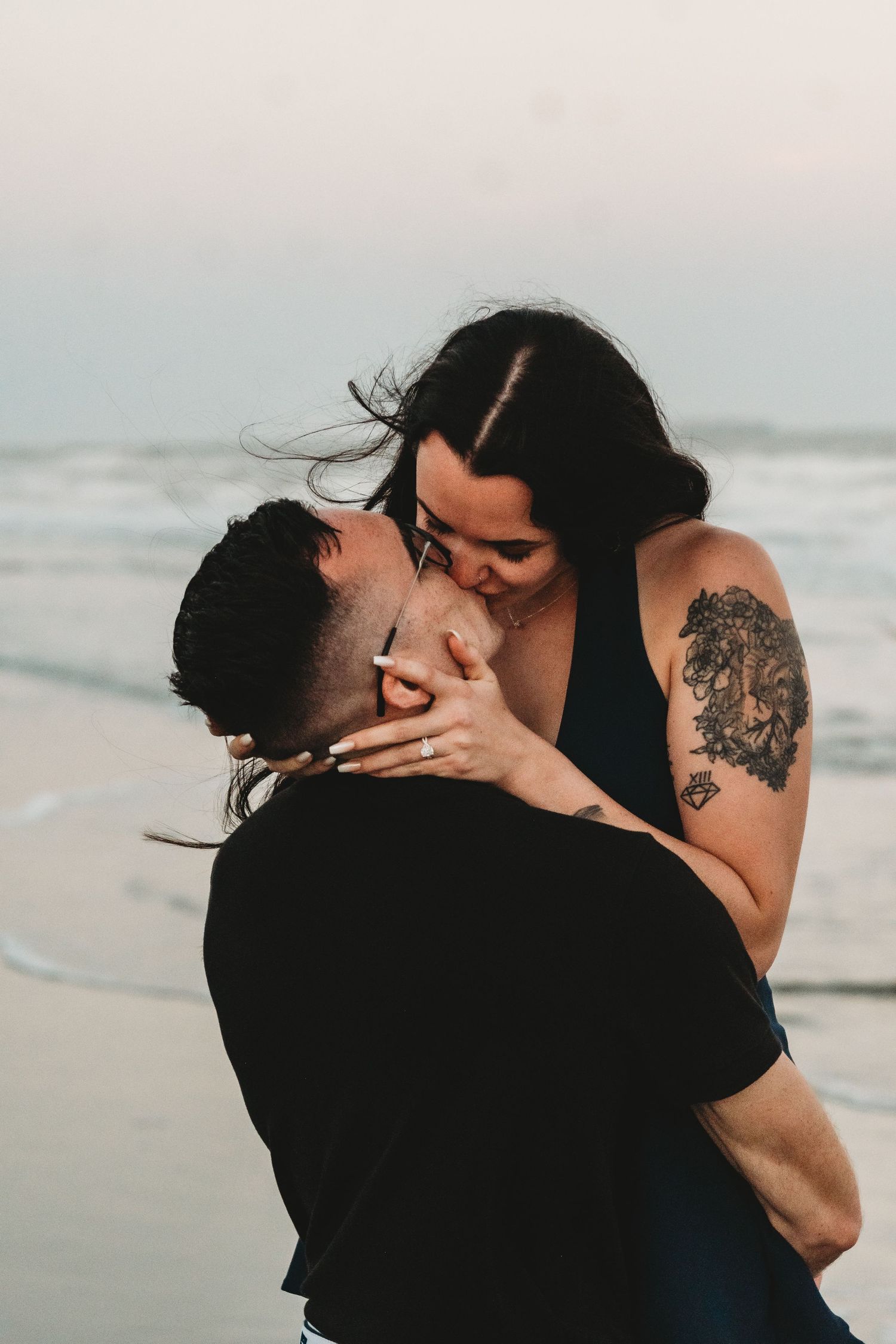 A couple shares a romantic kiss on a beach, woman with shoulder tattoo embracing man in black shirt by the ocean.