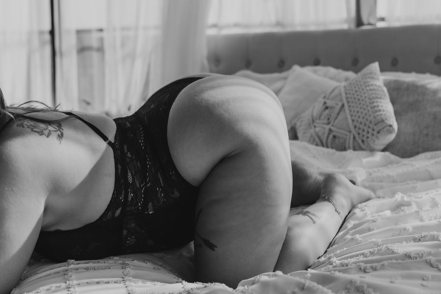Black and white boudoir photo of a woman in lace lingerie posing on a bed with soft natural lighting.