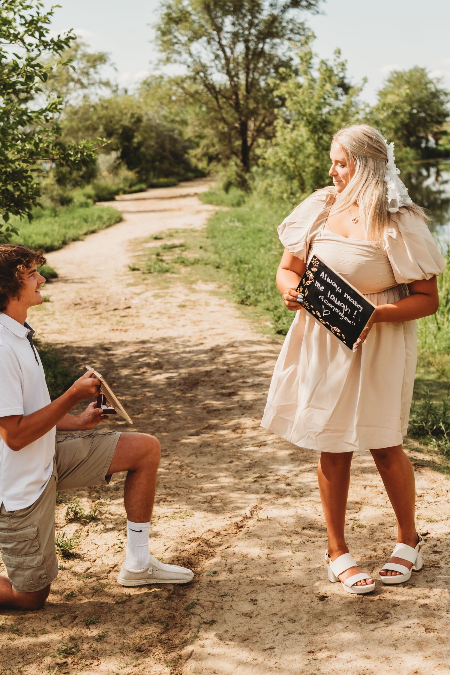 Man kneeling on sandy path proposing to blonde woman in beige dress holding chalkboard sign outdoors.