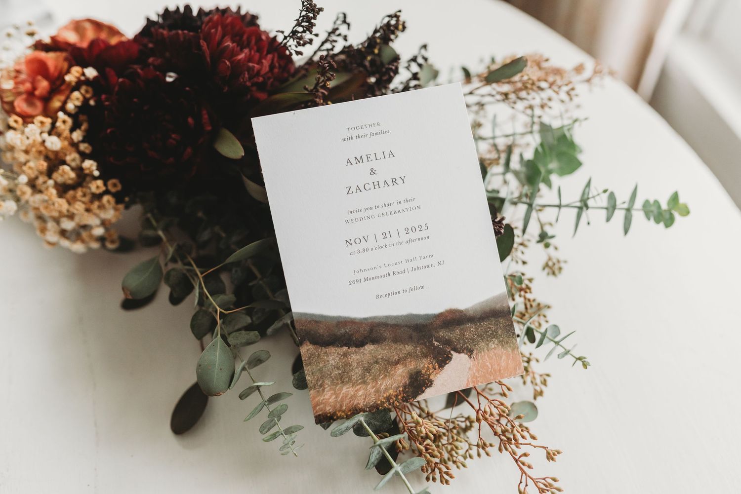 Elegant wedding invitation for Amelia and Zachary displayed among a rustic floral arrangement with burgundy and dried flowers.
