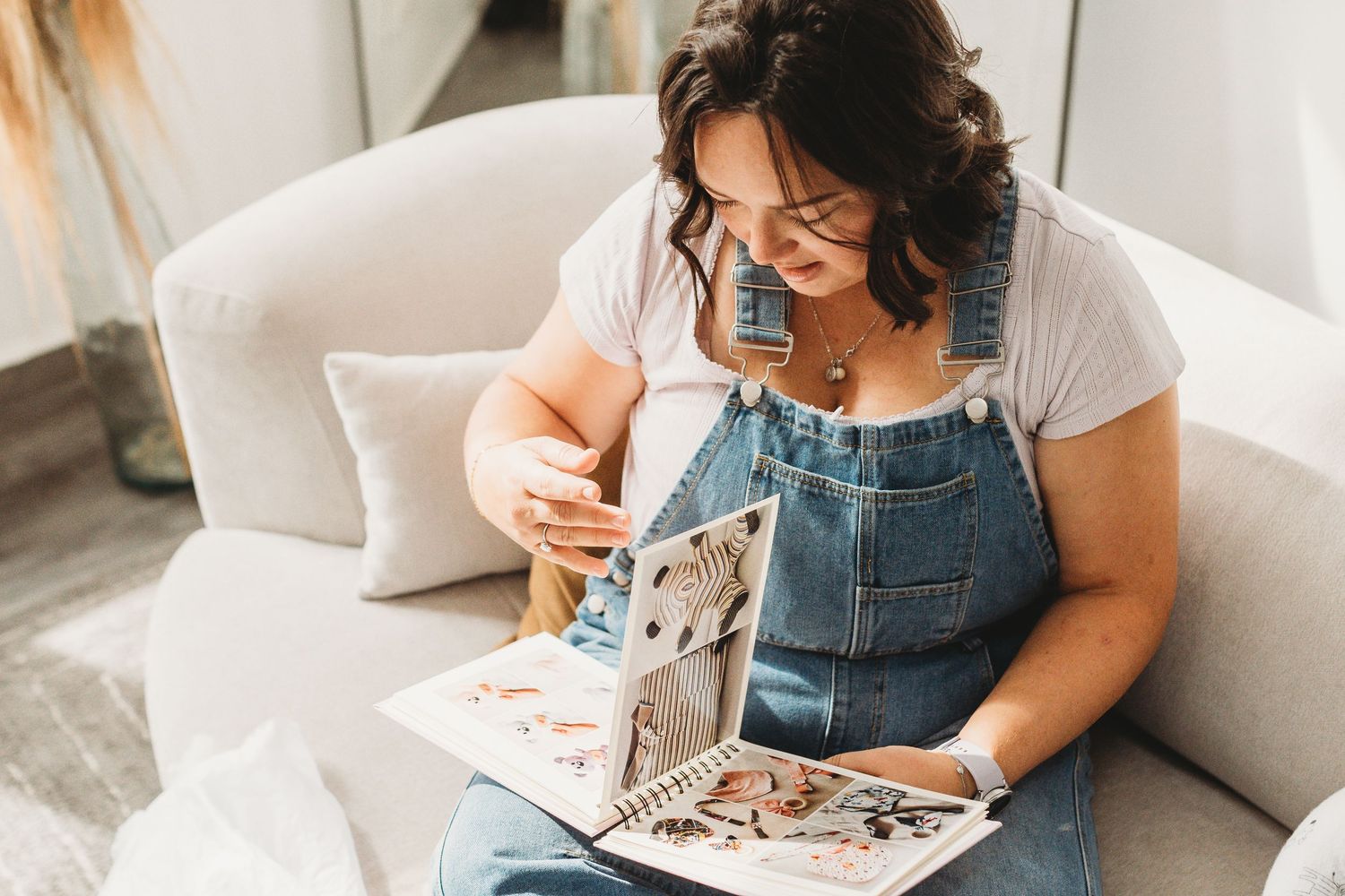 Person in denim overalls reading book while relaxing on white couch.
