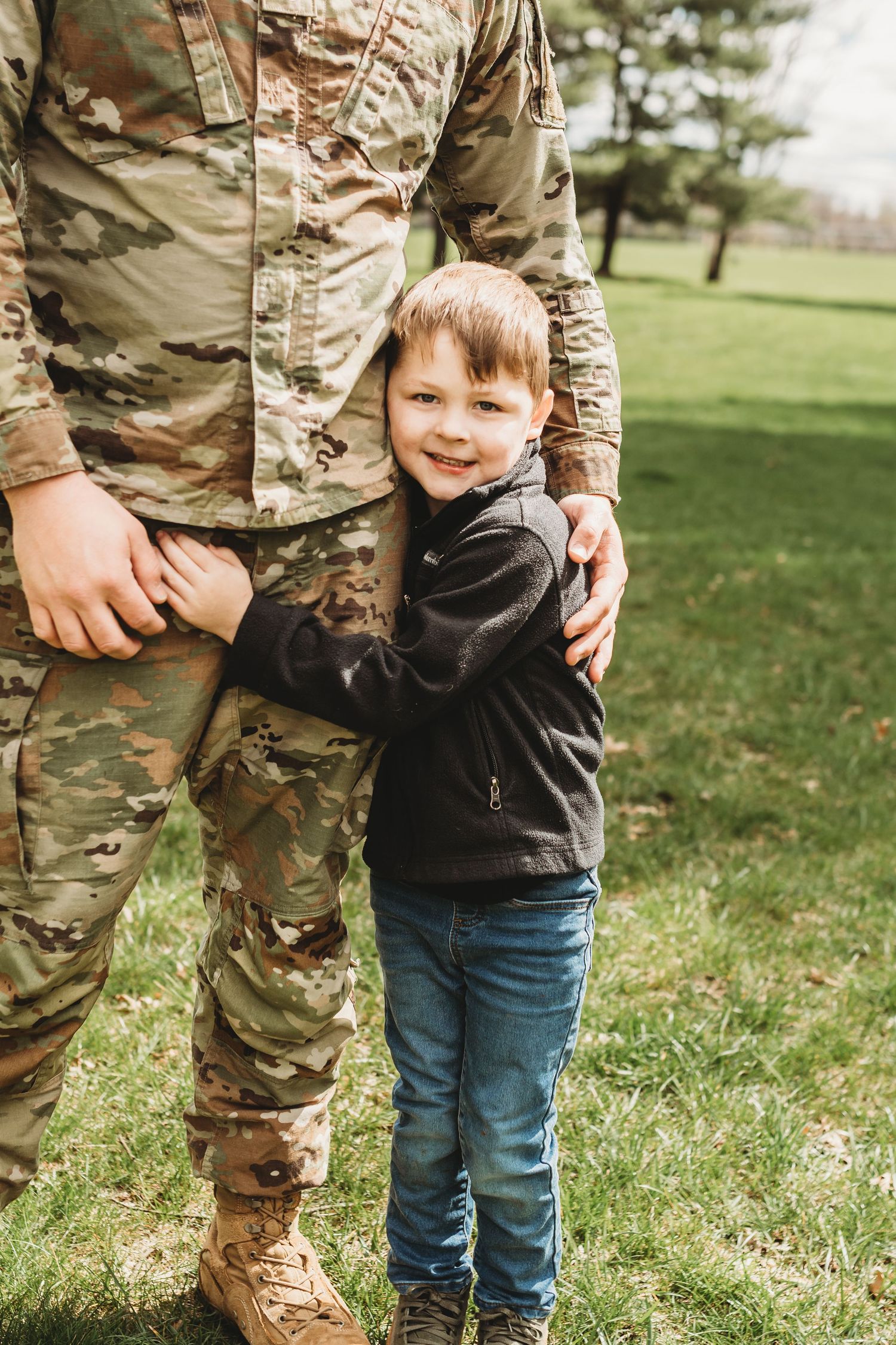 A small child stands close to someone wearing military camouflage fatigues on grassy outdoor terrain.