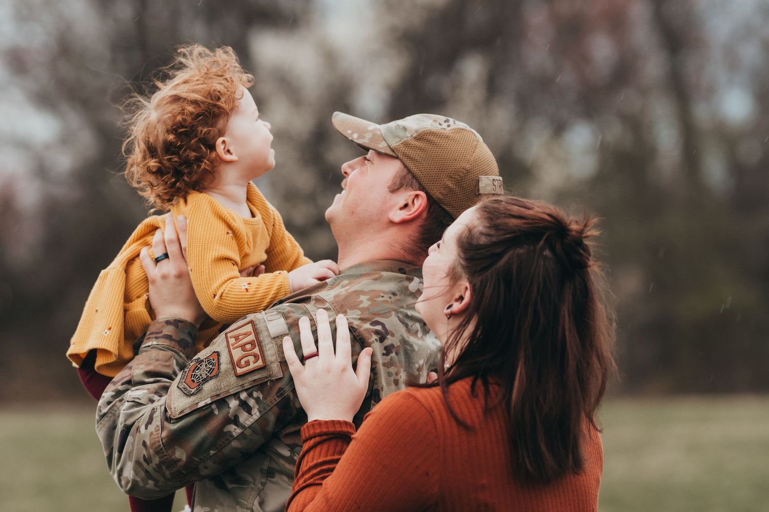A military service member in camouflage uniform shares a joyful moment outdoors with family members wearing orange sweaters.