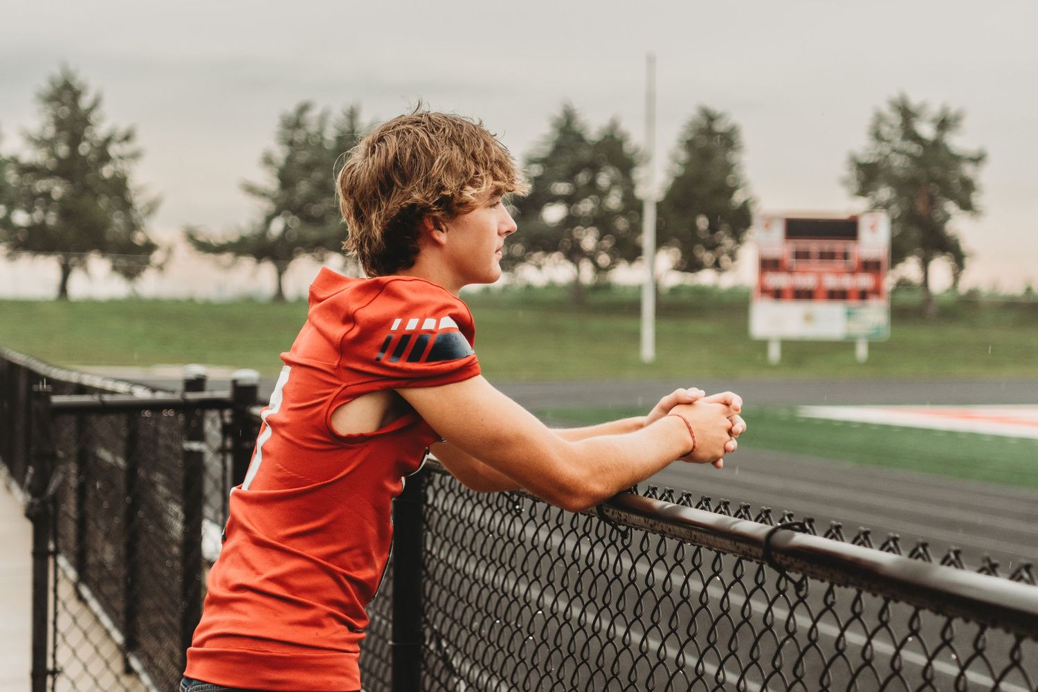 Baseball player in red uniform leans against chain link fence at high school sports field.