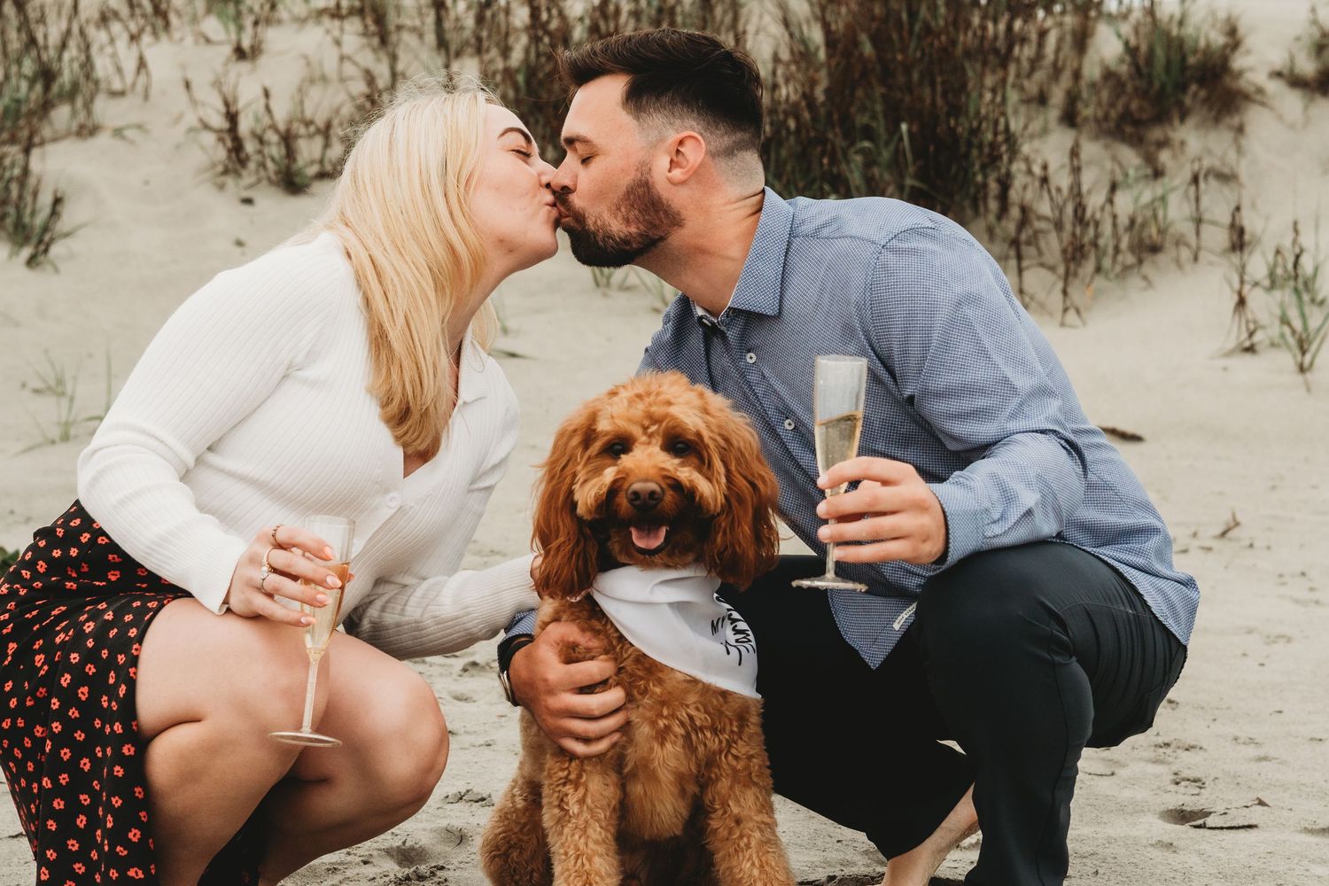 Couple kissing while crouching with their golden doodle dog on a sandy beach, man holding champagne glass.