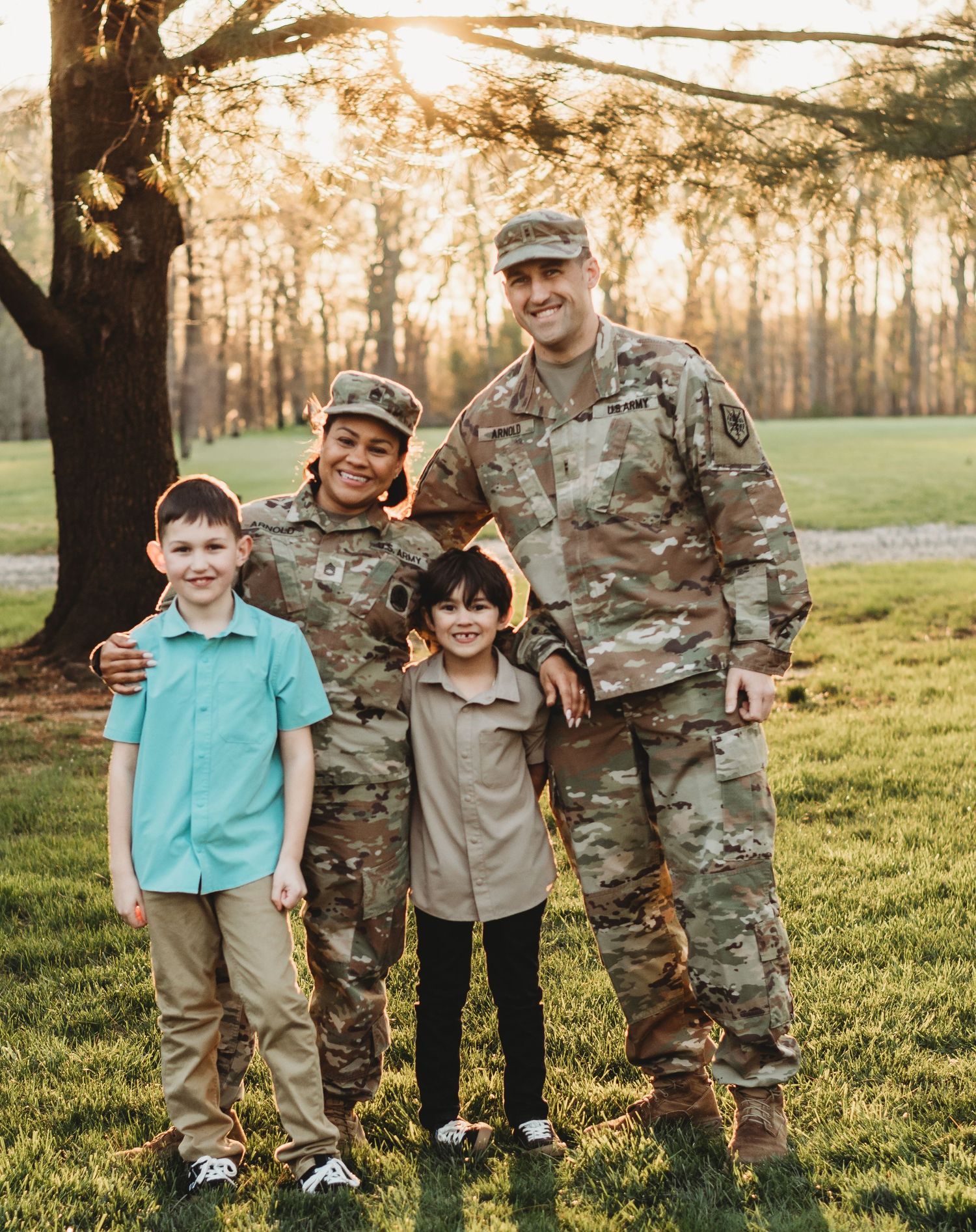 Military service members in camouflage uniforms pose with children on grassy field at sunset.