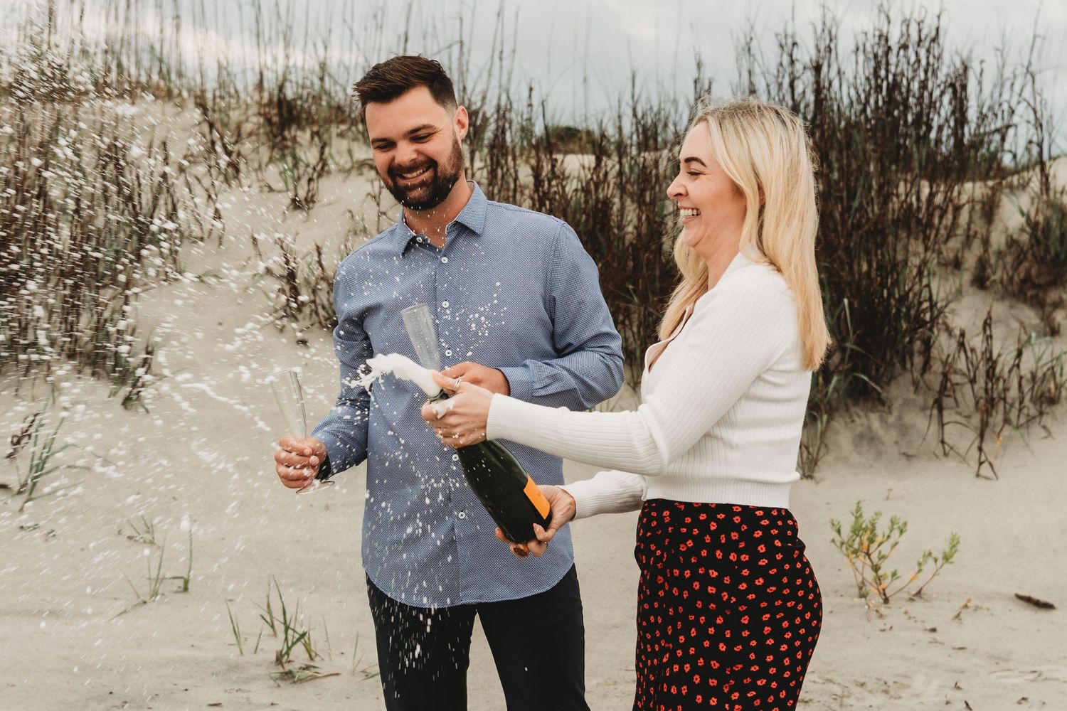 Couple celebrating on a sandy beach, popping champagne bottle together, smiling joyfully outdoors.