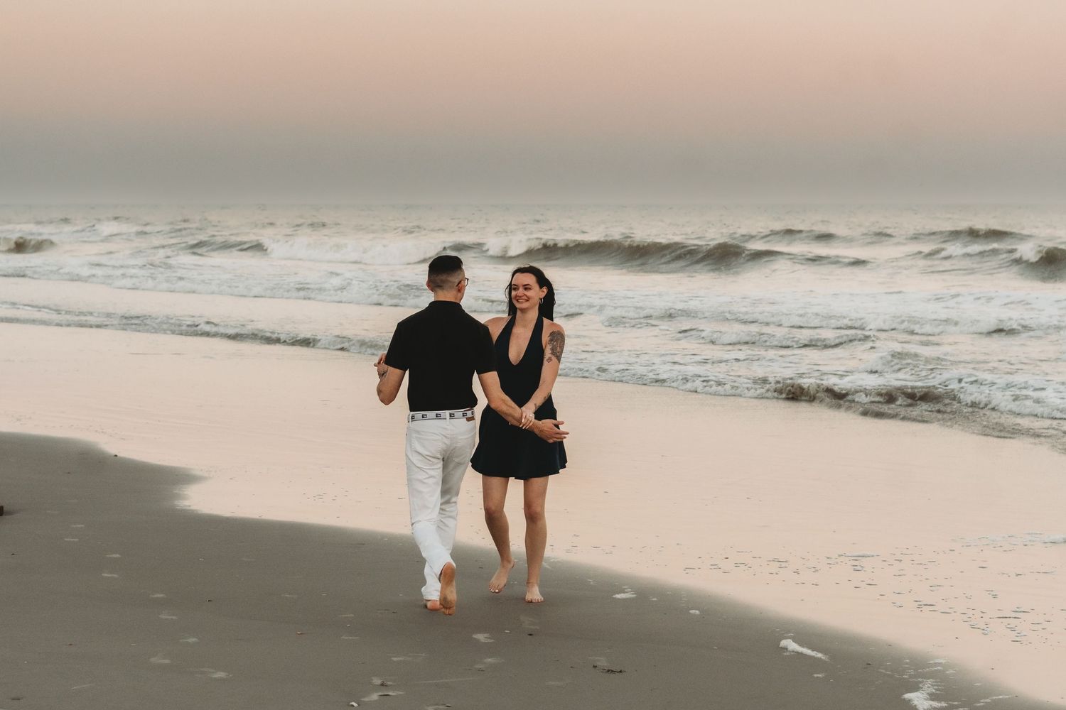 Couple in black outfits walking hand in hand on a sandy beach at sunset with ocean waves in the background.