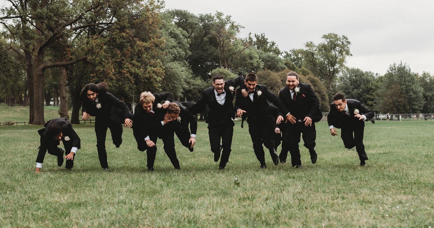 Groomsmen running playfully across a green lawn during wedding festivities.