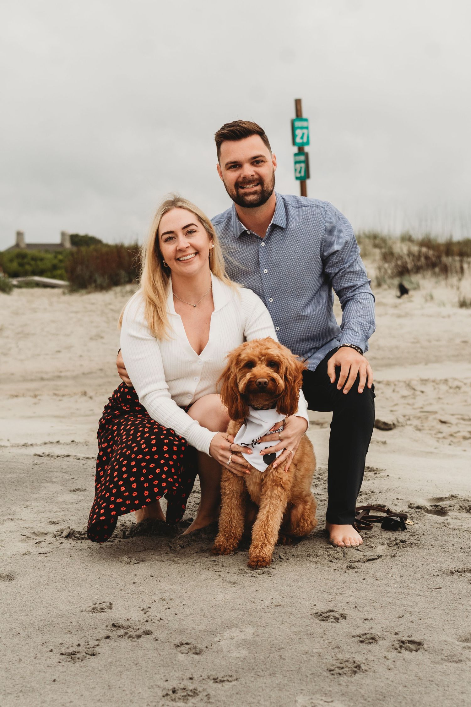 Smiling couple posing with a golden doodle dog on a sandy beach on a cloudy day.