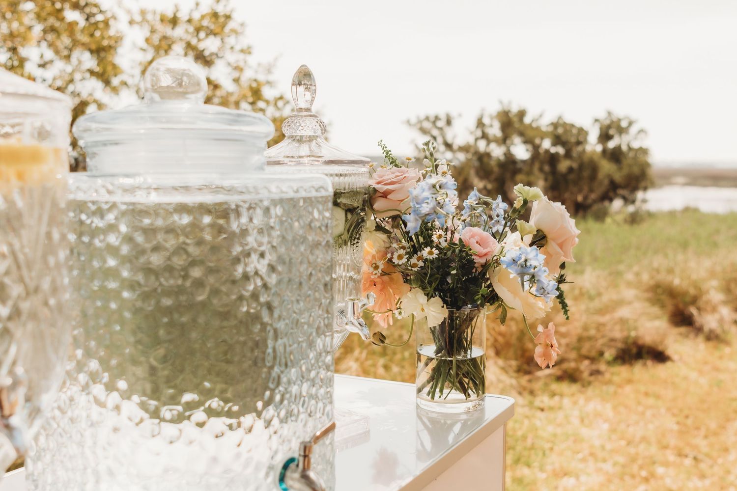 A glass beverage dispenser and floral bouquet arrangement sit outdoors against a sunlit natural landscape.