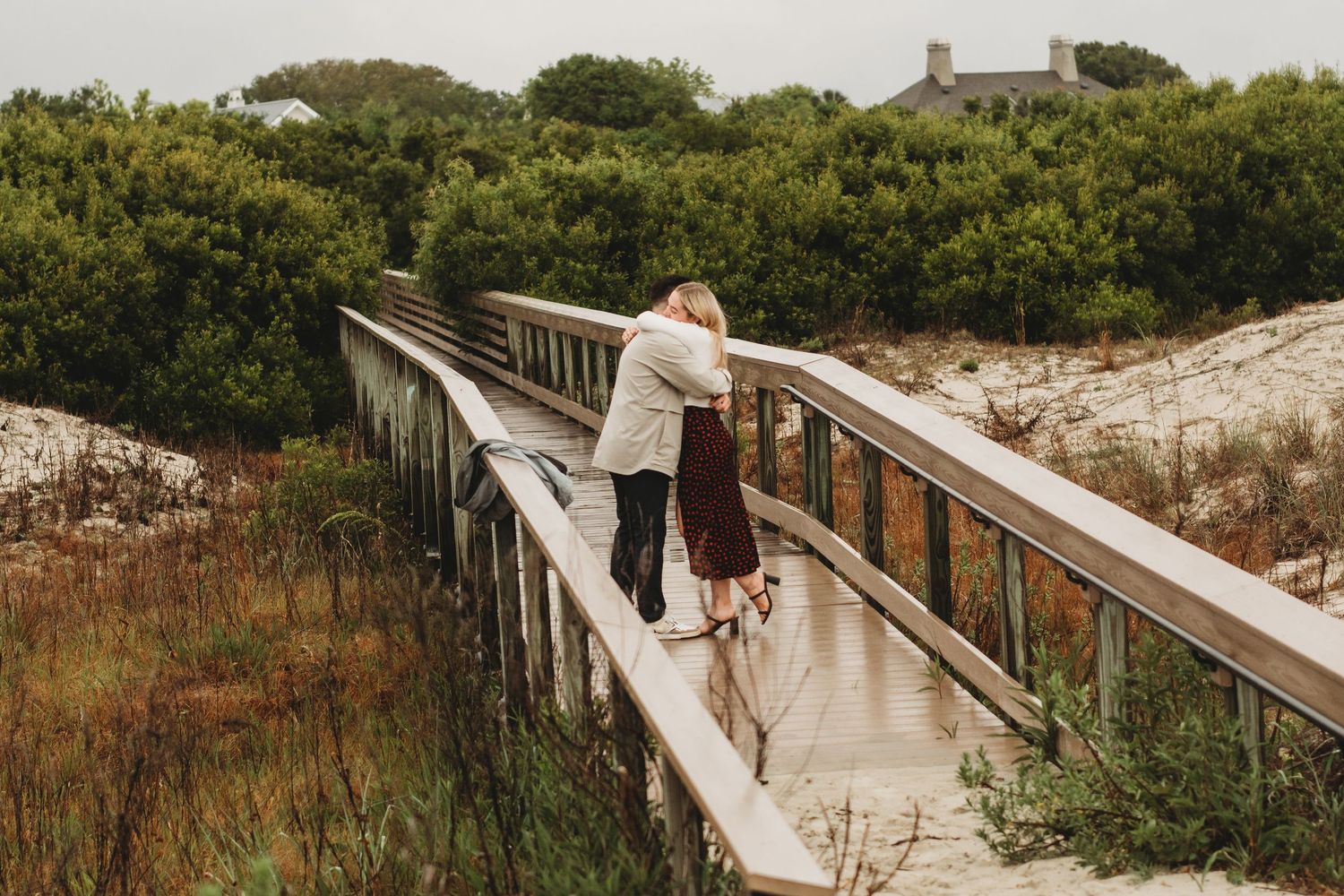 Couple sharing a romantic kiss on a wooden bridge surrounded by lush greenery and sandy dunes.