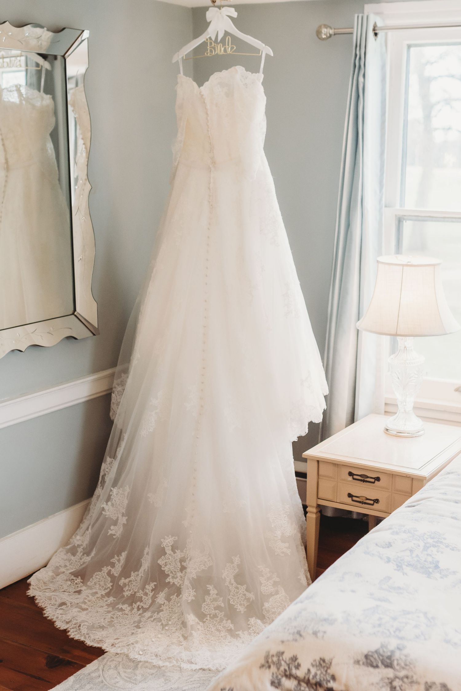Elegant white wedding gown with lace details displayed on a hanger against a light gray wall.