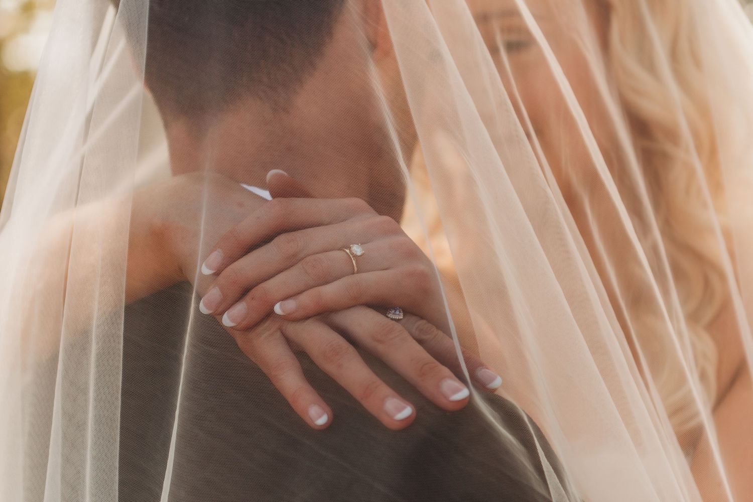 Bride and groom embrace under sheer wedding veil, showing diamond engagement ring and wedding band on bride's hand.