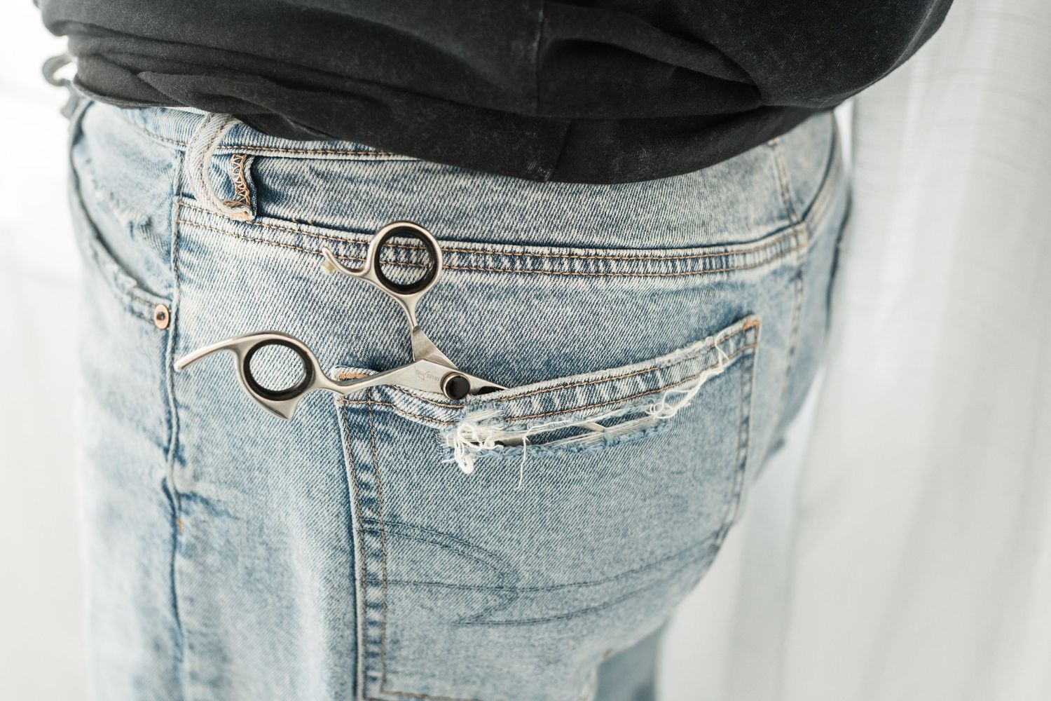 Close-up of light wash distressed jeans with scissors tucked into the front pocket, worn with a dark top.