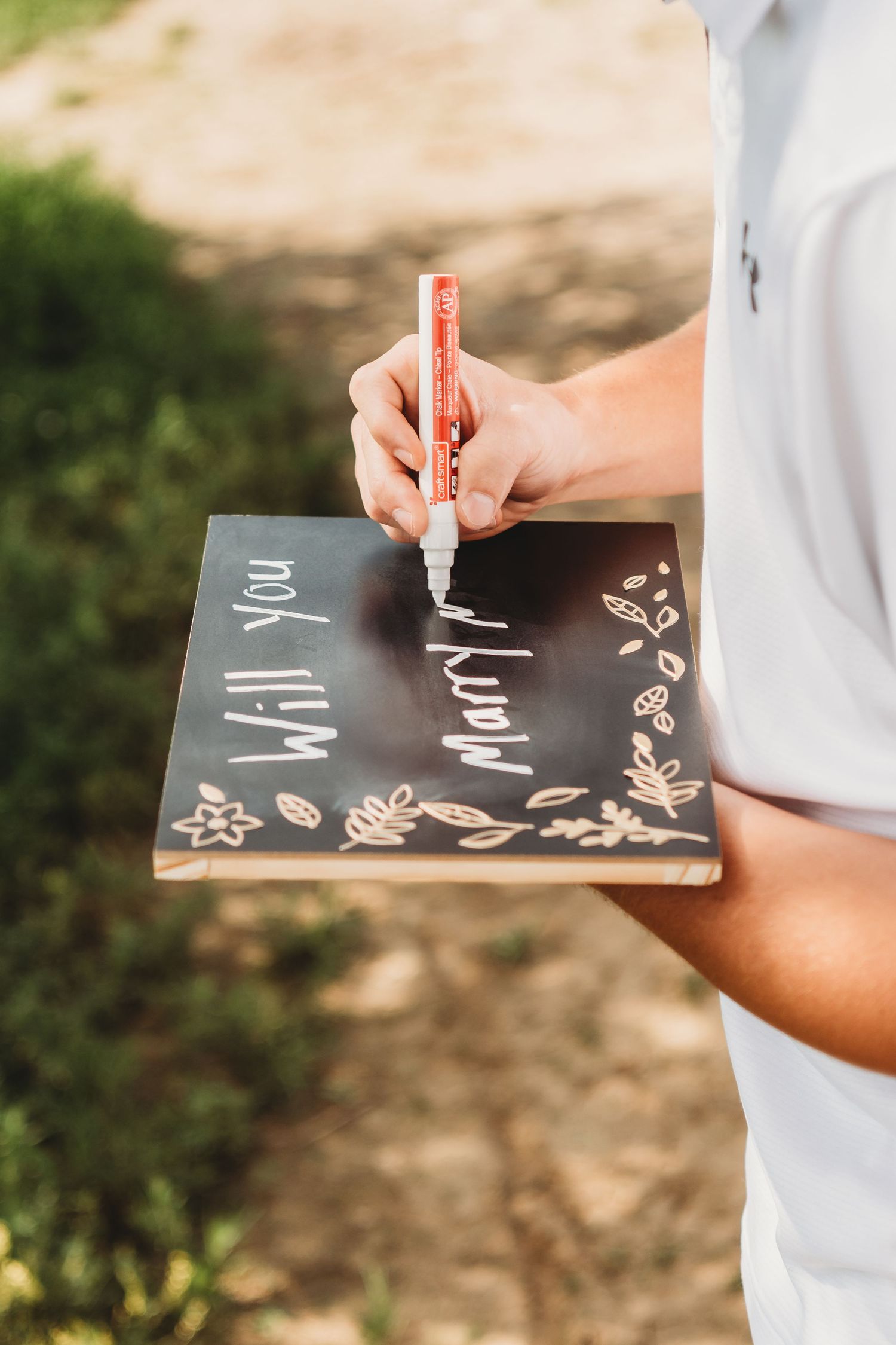 Person writing Will You Marry Me on a decorative black chalkboard sign with a white marker outdoors.