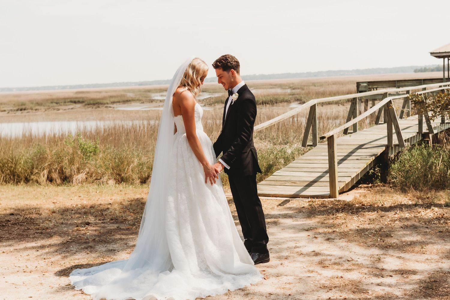 Bride and groom share a romantic moment near a wooden bridge overlooking coastal marshlands on their wedding day.