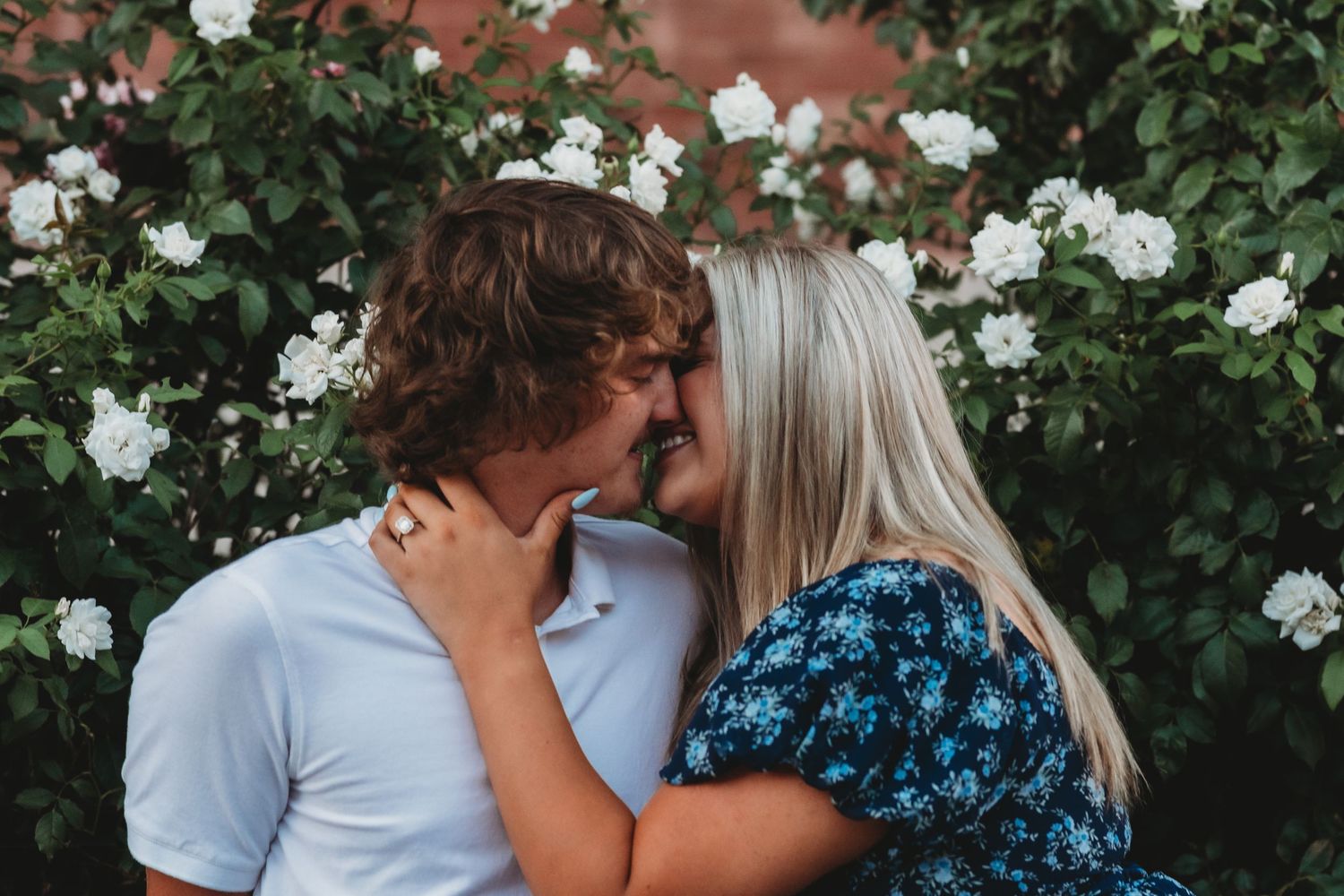 Couple sharing a romantic kiss in front of lush green bushes with white roses in an outdoor garden setting.