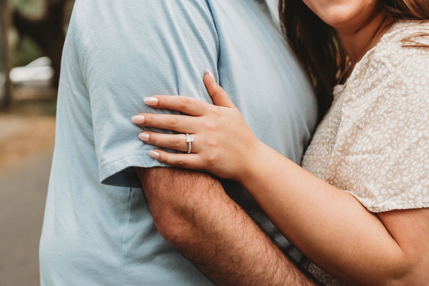 Close-up of a couple embracing, woman wearing a diamond engagement ring, dressed in casual summer outfits outdoors.