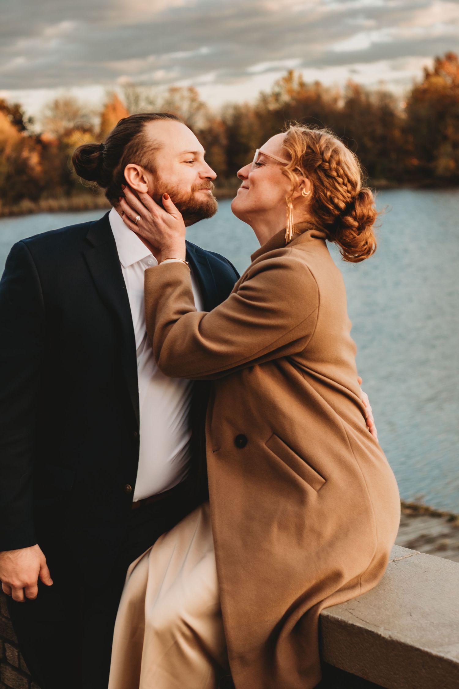 Romantic couple embracing by a lake during golden hour wearing formal attire.