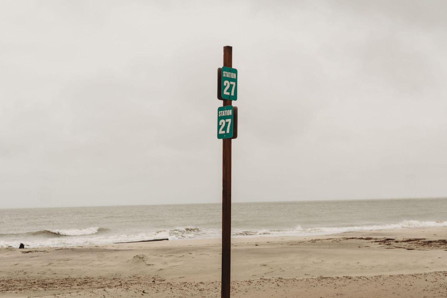 Green Route 27 signs on a wooden post at a sandy beach with overcast skies and ocean waves.