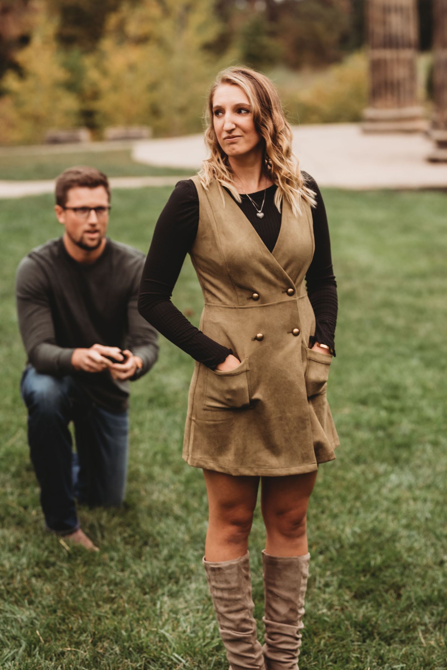 Woman in olive vest dress and knee-high boots stands on grass, man kneeling with phone in background.