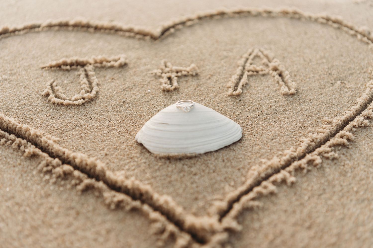 Heart drawn in sand with initials J+A and a white seashell, romantic beach love symbol.