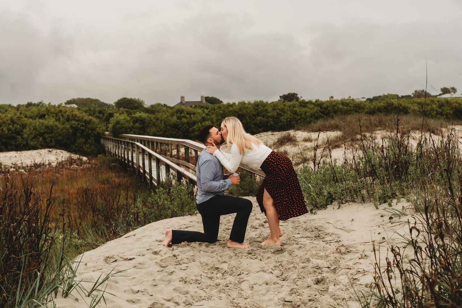 Man proposes on sandy beach dunes while couple kisses near wooden boardwalk with lush greenery and cloudy sky.