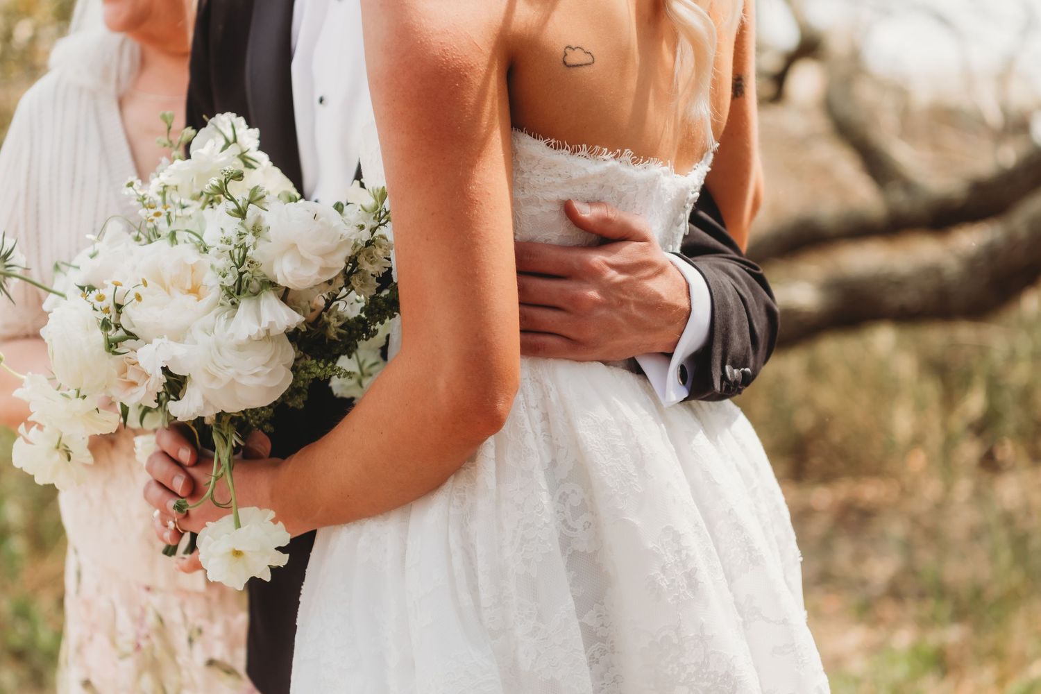 Bride and groom embracing outdoors, bride holding white floral bouquet in elegant lace wedding dress.