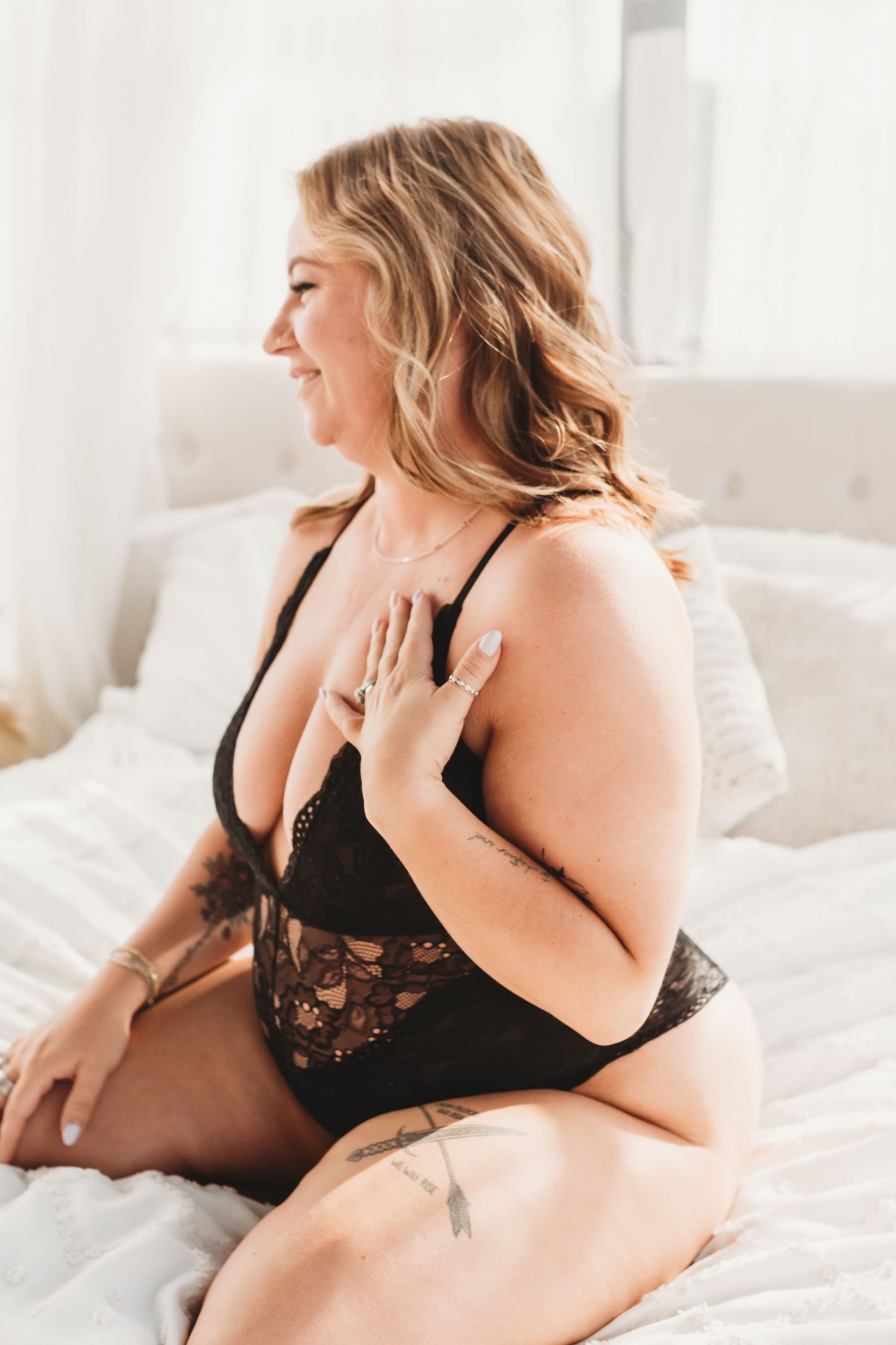A smiling plus-size woman in black lace lingerie sits on a white bed, showcasing a boudoir photography style.