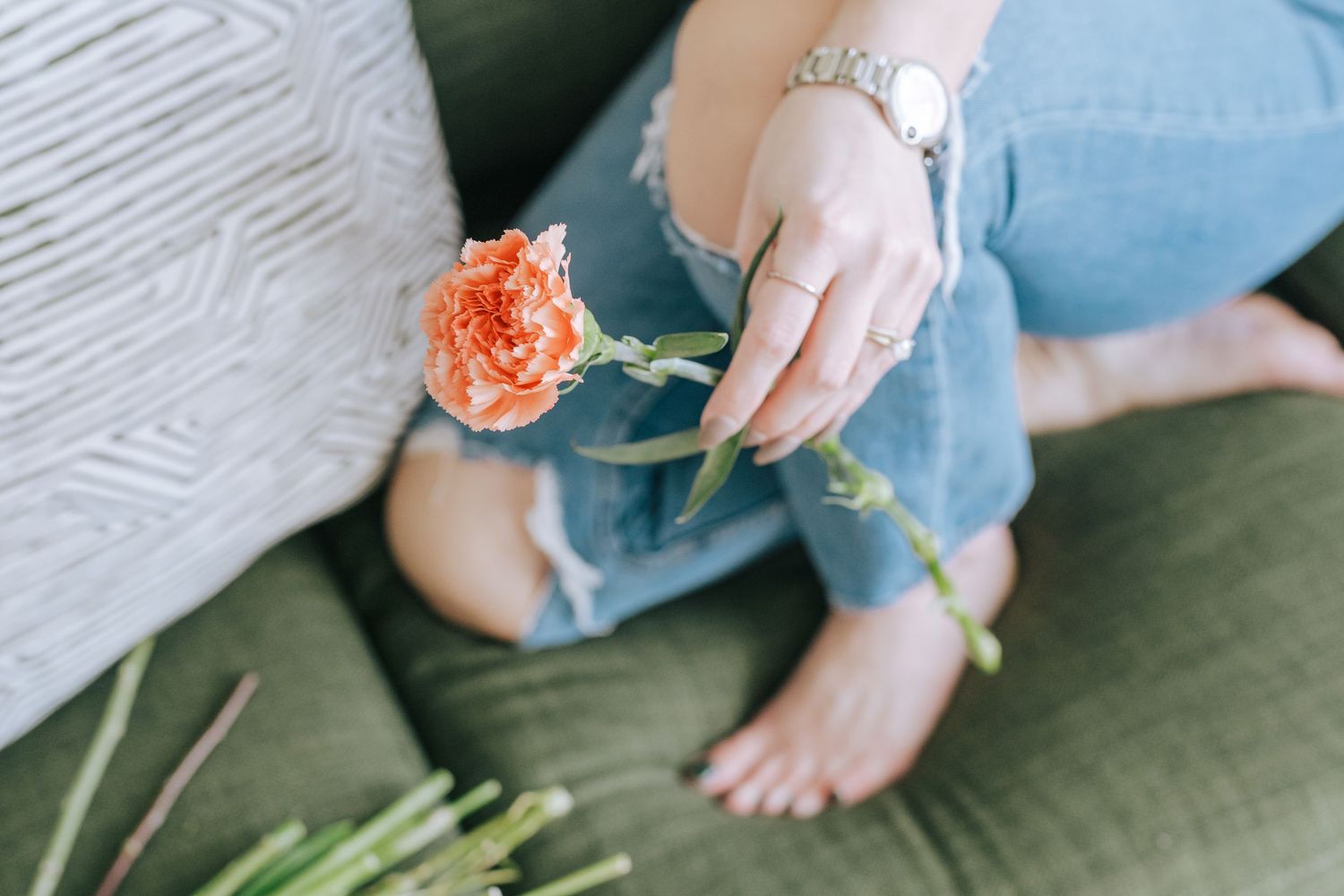 Single orange carnation held by hand with painted nails against green cushion.