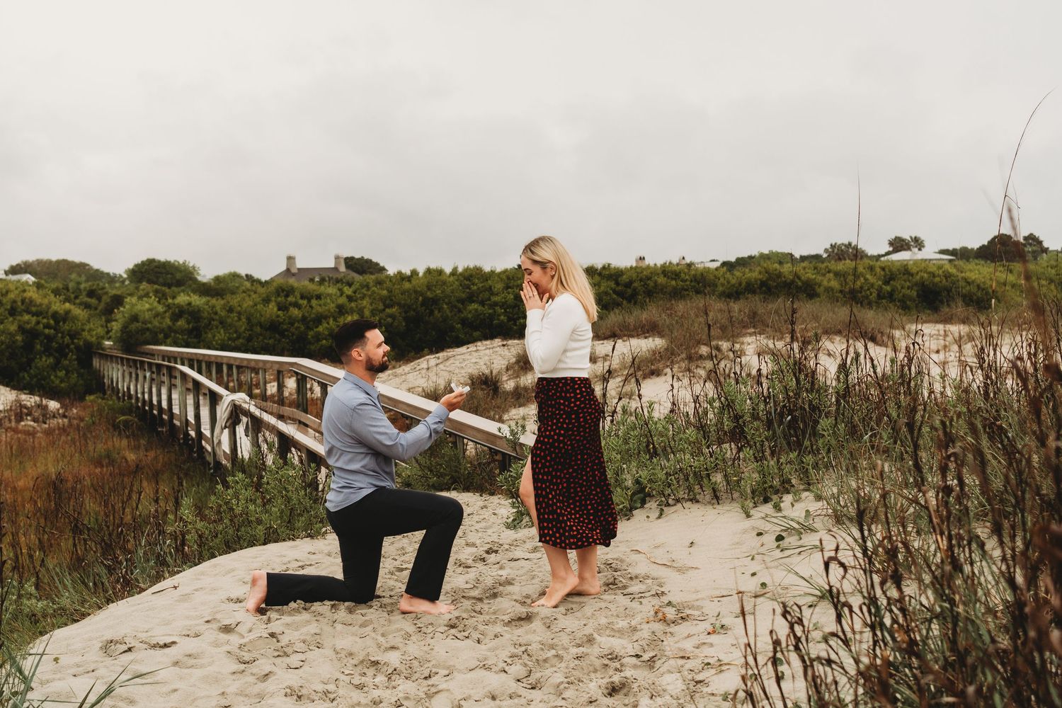 Man proposing on one knee to woman on sandy beach path with bridge and greenery backdrop.