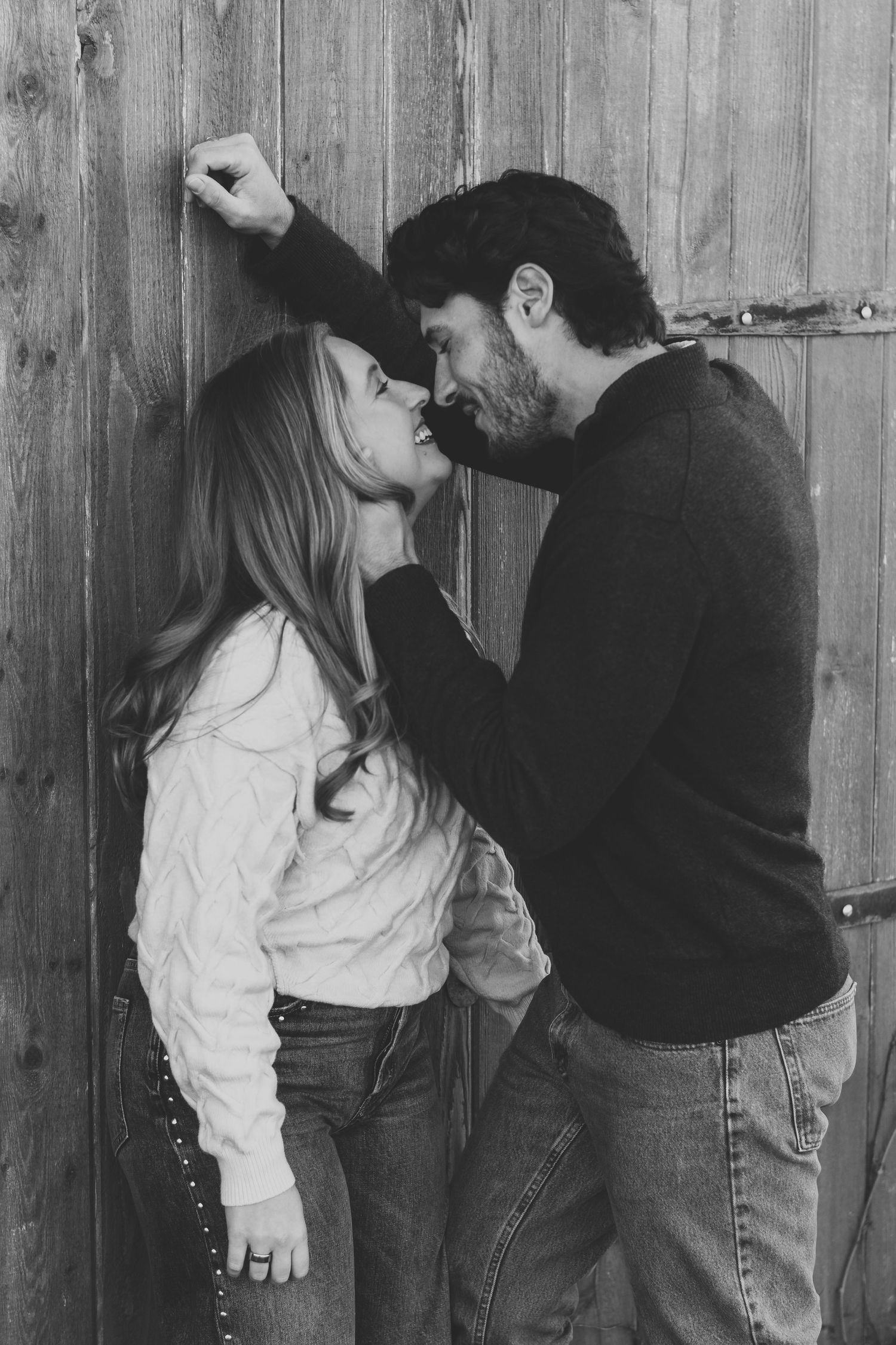 Black and white photo of a couple embracing against a rustic wooden wall.