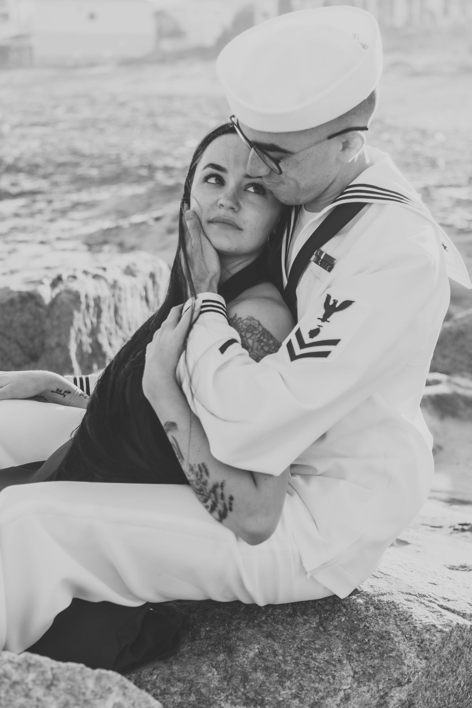 Black and white photo of a sailor in uniform tenderly embracing a woman on rocky shore.