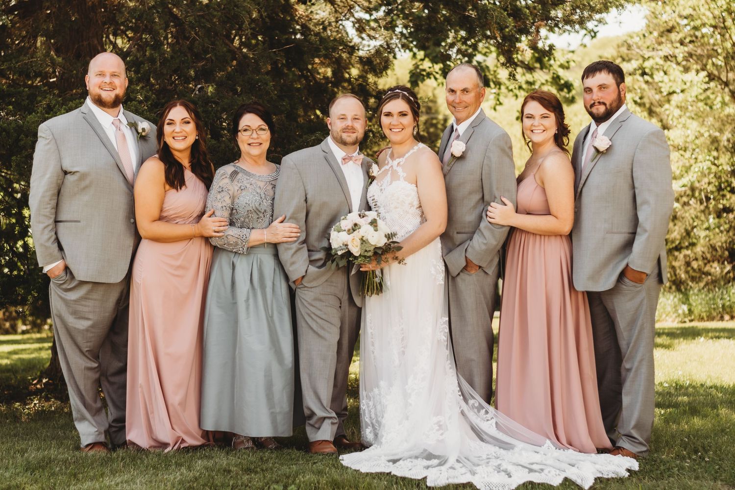 Wedding party posed outdoors wearing pink and grey attire against autumn foliage.