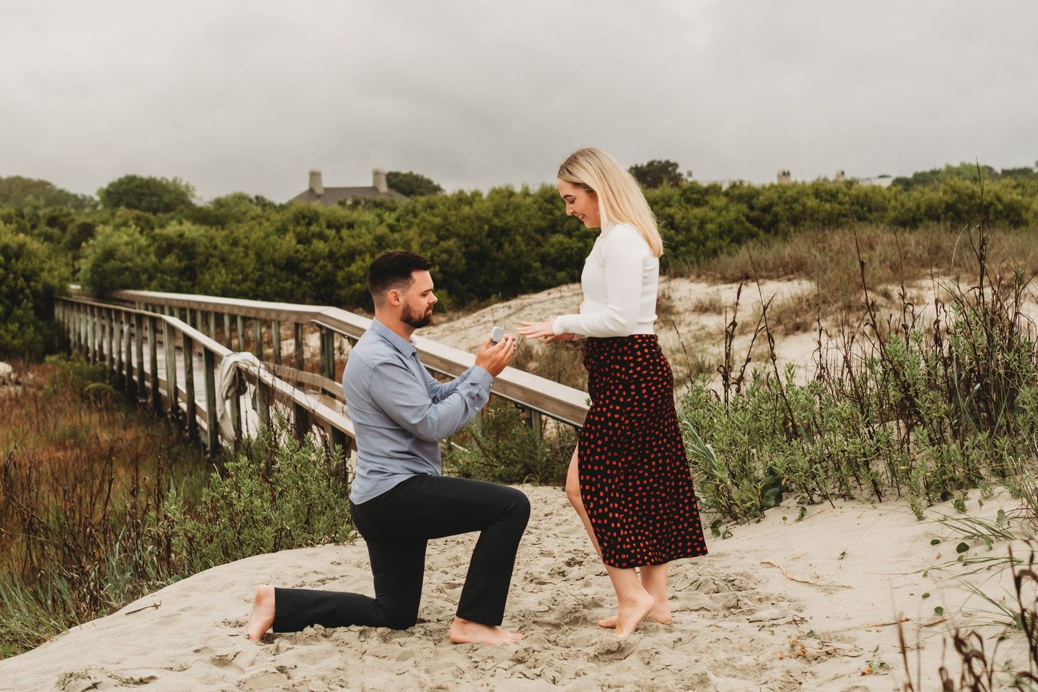Man proposing on one knee to woman on sandy beach near wooden bridge, romantic outdoor engagement scene.