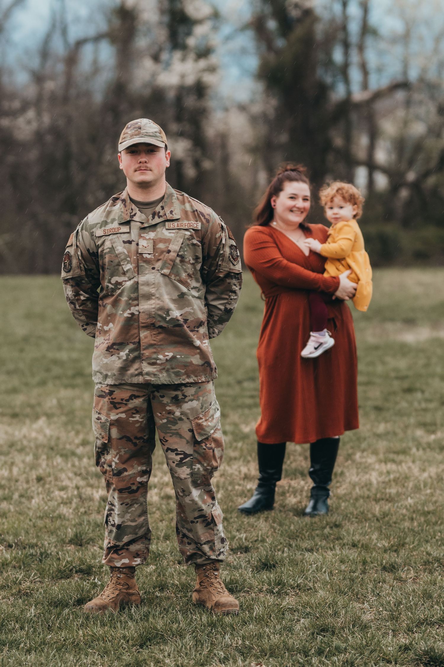 Military service member stands in uniform on grassy field while family members gather in background.