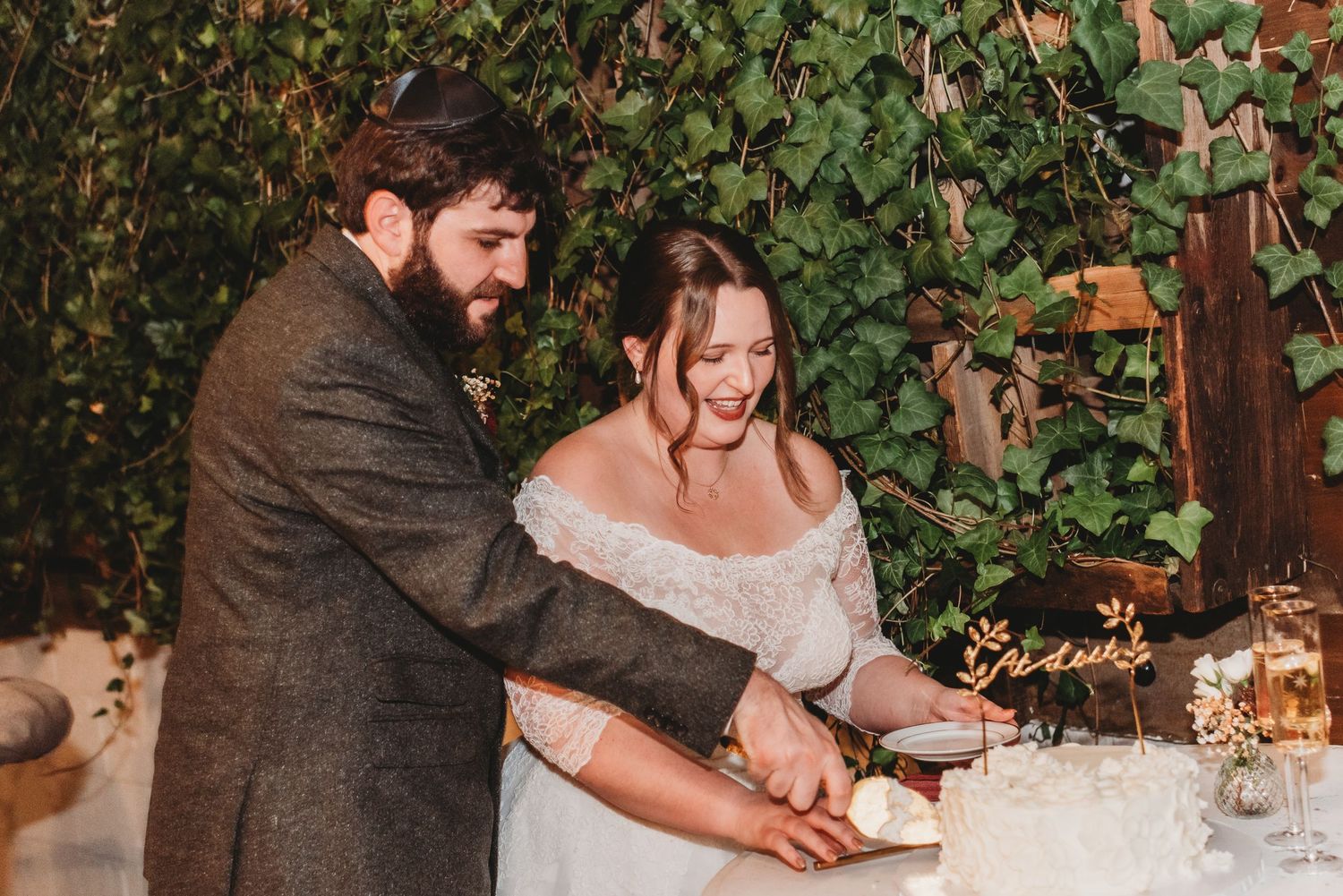 Bride and groom smiling while cutting their wedding cake together at an elegant outdoor reception with ivy backdrop.