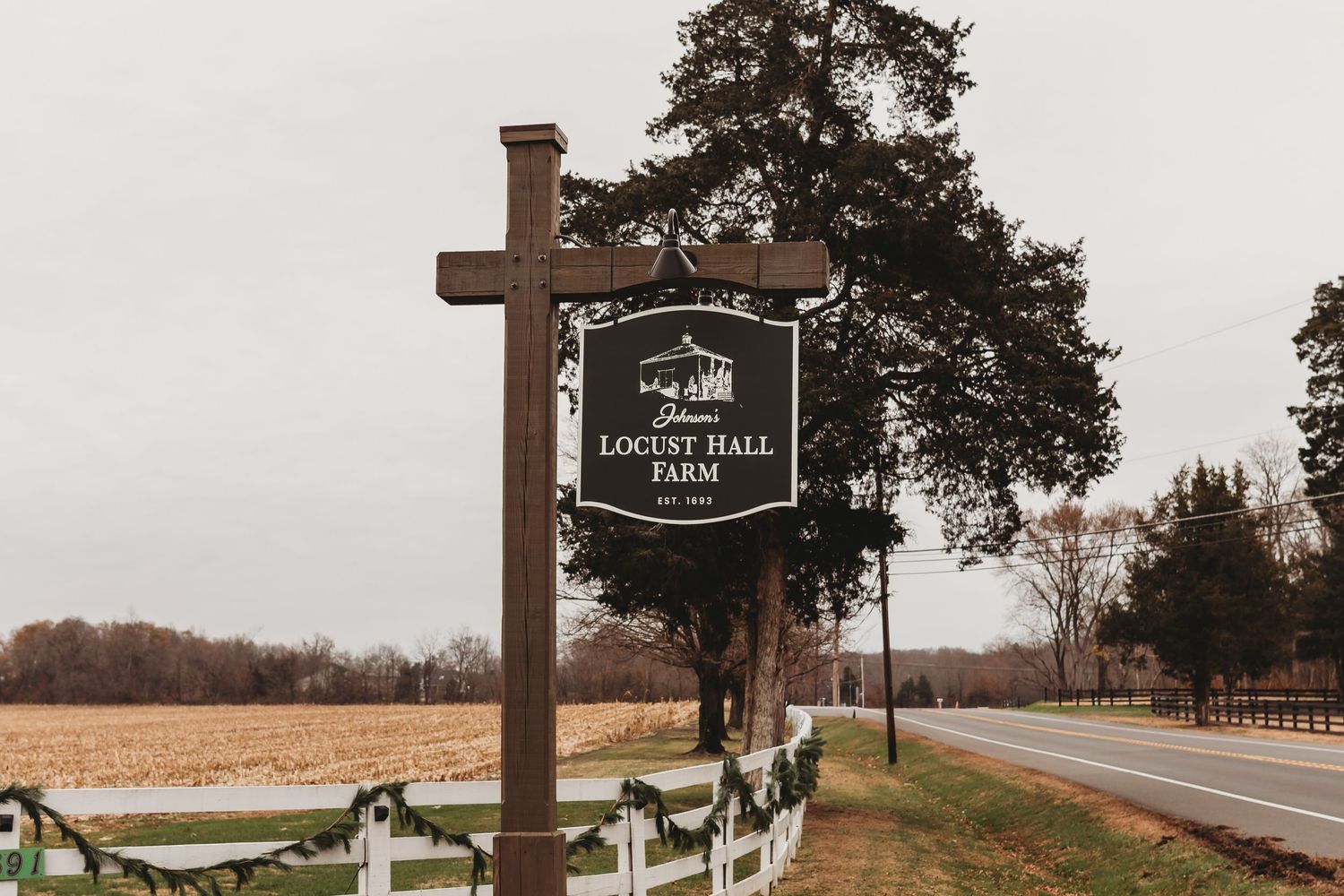 Rustic wooden sign for Locust Hall Farm along a scenic rural road with white fence and bare trees.
