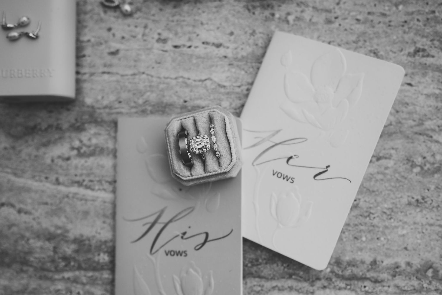 Black and white photo of wedding rings in a box beside His and Her vow books on a marble surface.