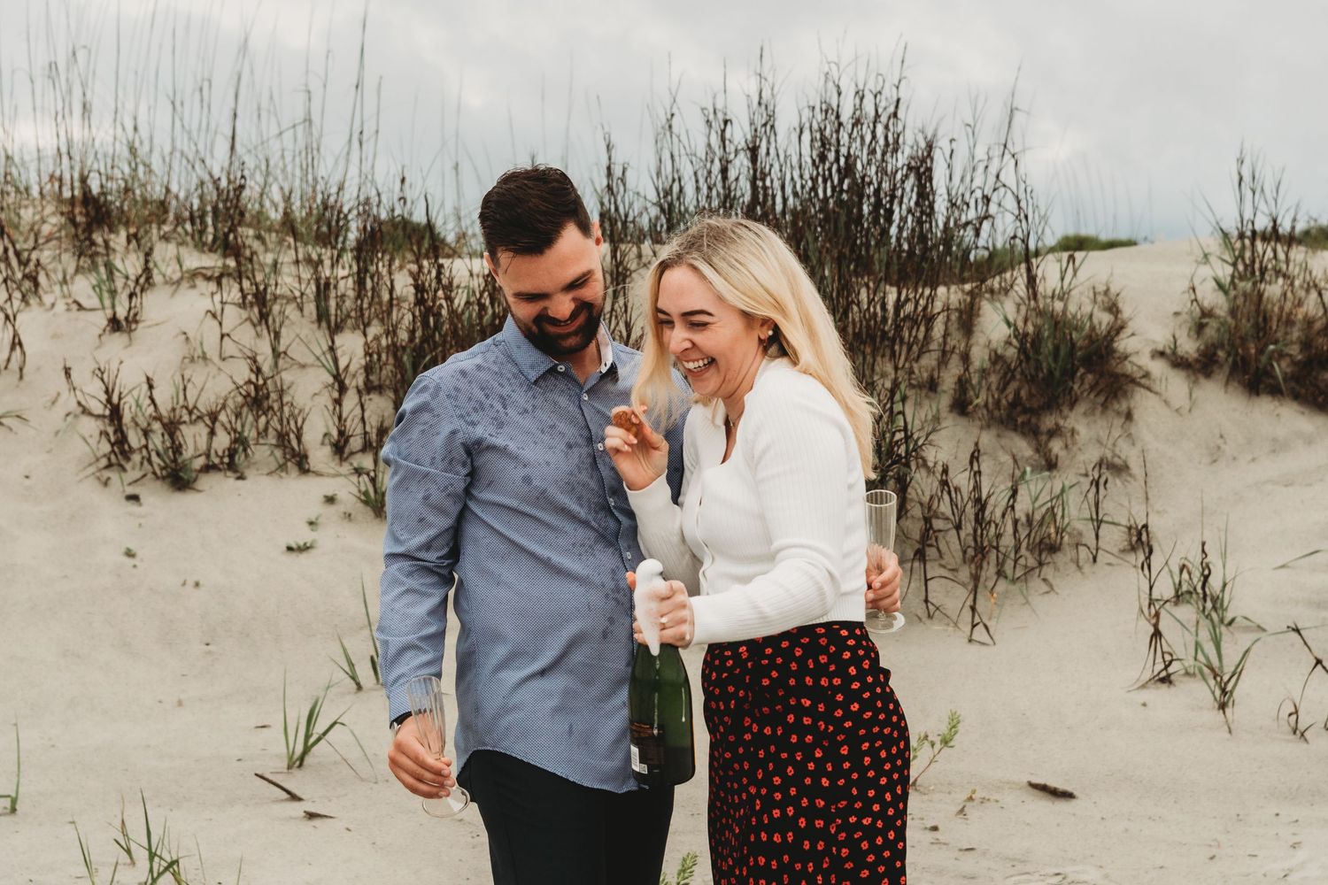 Happy couple celebrating on sandy dunes, woman holding champagne bottle, laughing together outdoors.